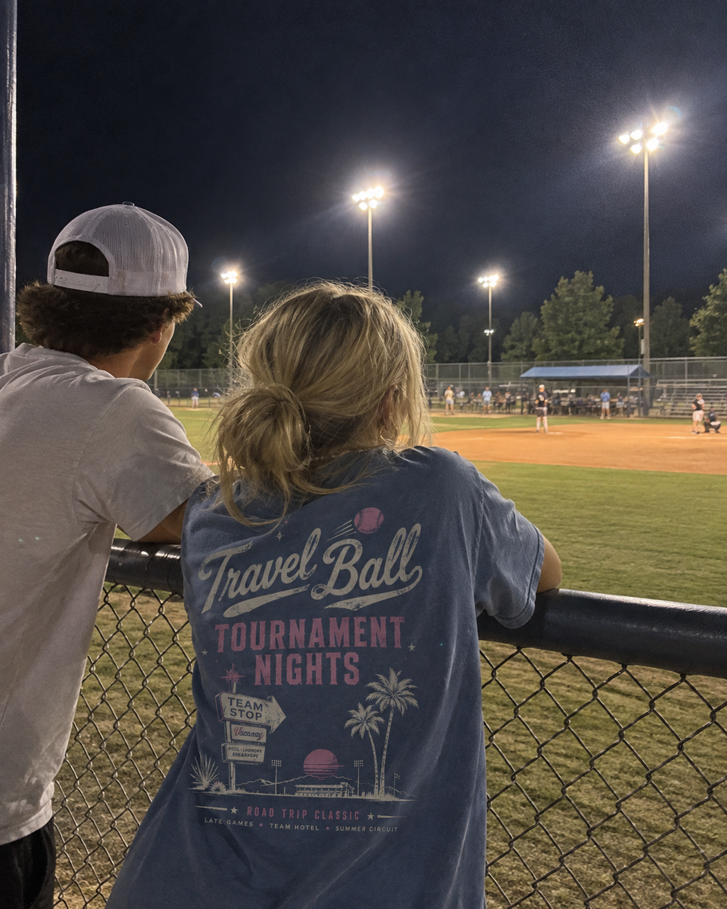 Teen girl wearing the Washed Denim adult baseball T-shirt at a ballpark, showing the large off-white and pink Travel Ball back graphic while watching a baseball game.
