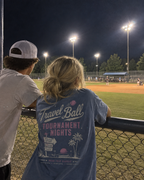 Teen girl wearing the Washed Denim adult baseball T-shirt at a ballpark, showing the large off-white and pink Travel Ball back graphic while watching a baseball game.